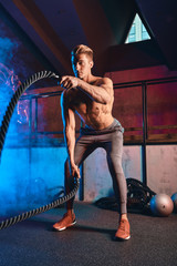 Shirtless strong athlete being concentrated on the exercise with battle rope requiring increased effort and strength. Cross Fit concept.