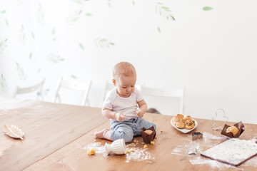 Happy infant boy eats ready-made cakes and having fun with cutting board covered with flour after cake dough sitting on wooden kitchen table. Child s development in home interior