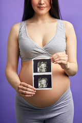 Unrecognizable pregnant white skinned woman in grey home suit with top holding ultrasonic scan in...