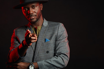 Black jazz musician man wearing fashionable suit and grey hat aiming at camera with one hand