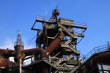 Fototapeta na wymiar Landschaftspark Duisburg, Germany: Low angle view on stairways into deep blue sky at corroded tower with rusty pipeline