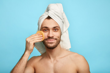 good looking pleasant man wiping face with cosmetic sponge, isolated over blue background, copy space