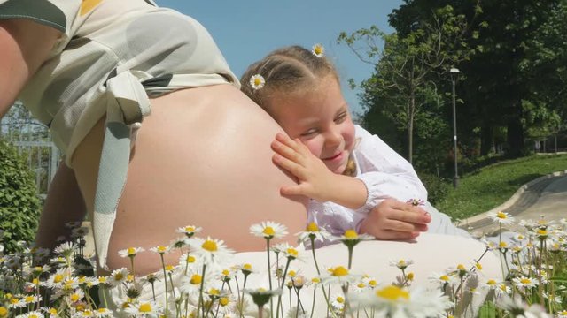 Young mother who pregnant for second time among the flowers with her daughter have fun outdoors, happy little girl leaned over mother's belly
