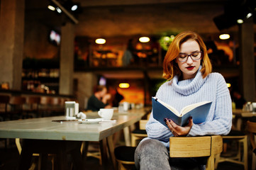 Cheerful young beautiful redhaired woman in glasses sitting at her working place on cafe reading something at her notebook.