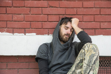 Homeless man, Homeless man drug and alcohol addict sitting alone and depressed on the street leaning against brick building wall feeling anxious and lonely, social documentary concept hard contrast 