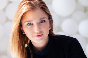 Fototapeta premium Cute beautiful pretty attractive romantic girl in black sweater looking at camera, female beauty portrait with makeup in rays of sun against background of white balloons in photostudio