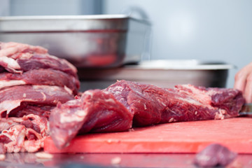 Closeup hands of man butcher chef cutting slices of fillet raw beef and pork meat off loin with knife in restaurant on red plastic board. Concept carve meat for barbeque grill on farmer market