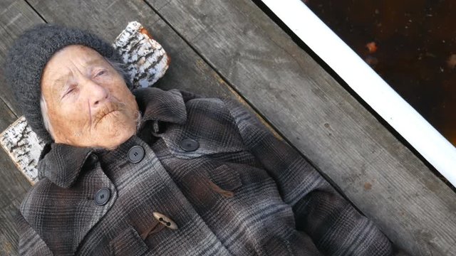 Grandmother lies on the street on wooden bed and looks at sky. Clouse up.  Pan from rotten apples on an old woman. Grandmother is wearing a coat and hat. She looks to the top. It's cold outside.