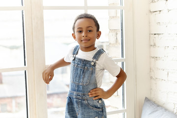 Portrait of confident handsome dark skinned African boy with short black curly hair standing by window at white brick wall, holding hand on his waist, going to have walk outside, looking at camera