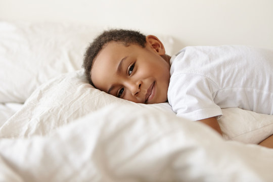 Coziness, happy childhood, relaxation and sleep concept. Picture of adorable charming black dark skinned ten year old boy lazing in bed, lying on white soft pillow, smiling joyfully at camera