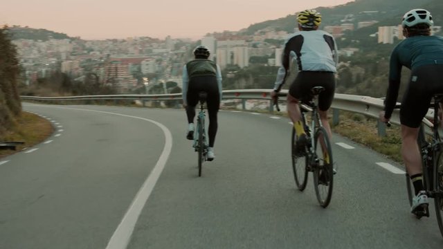 Three road cyclists ride down mountain in line formation, drafting in aero dynamic positions to save energy and speed. Beautiful sunset over city landscape in background