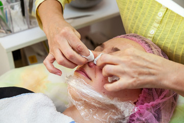 A young girl is lying on a couch during cosmetic procedures with a mask on the face above which beautician woman squeezes body fat and pimples with wipes on nose. Cleansing and make up.