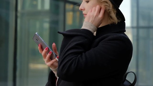 nice blond girl in black coat and hat listens to music with smartphone headphones against city street slow motion