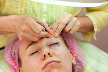 Obraz na płótnie Canvas A young girl is lying on a couch during cosmetic procedures with a mask on the face which beautician woman squeezes body fat and pimples piercing the skin with a needle on eye. Cleansing and make up.