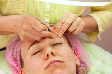 Obraz na płótnie Canvas A young girl is lying on a couch during cosmetic procedures with a mask on the face which beautician woman squeezes body fat and pimples piercing the skin with a needle on eye. Cleansing and make up.