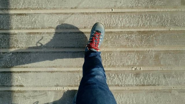 POV view from eyes to down on man walking in his big chunky leather blue boots with red laces on empty beach boardwalk covered with sand. Walking alone towards goals and success