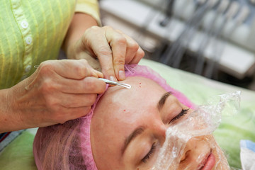 A young girl is lying on a couch during cosmetic procedures with a mask on the face above which beautician woman squeezes body fat and pimples with special metal tool in the form of blade on forehead