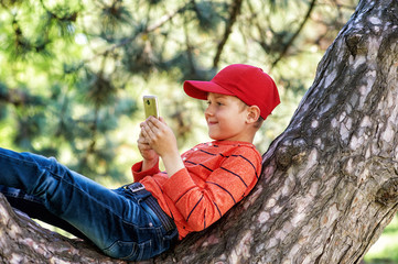 Cheerful boy sitting on a tree in the city Park . The child uses a mobile phone