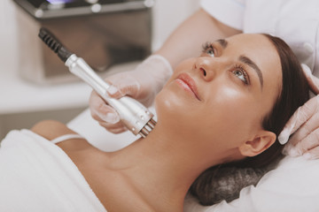 Beautiful woman getting microcurrent therapy session for her neck and face skin. Attractive woman smiling, getting skincare treatment at cosmetology clinic. Beauty, rejuvenation concept