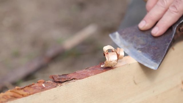 Man works as a hand tool hatchet, processes a board of pine tree by clearing it from the bark. Construction worker planing a piece of wood for a building project.