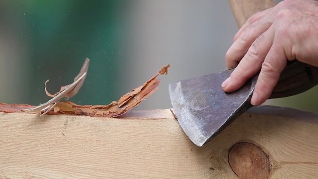 Man works as a hand tool hatchet, processes a board of pine tree by clearing it from the bark. Construction worker planing a piece of wood for a building project.