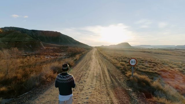 Aerial footage of drone flying back and revealing amazing dusty gravel road with a lonely solo figure of hipster traveller man, or drone pilot, look into distance at sunset or sunrise