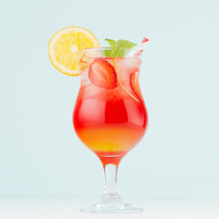 Tropical refreshing striped fruit drink sunrise with slices oranges, ice, strawberry, mint in wineglass on light pastel green interior, square.