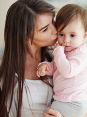 Beautiful young mother holding and kissing thoughtful baby daughter sucking her thumb