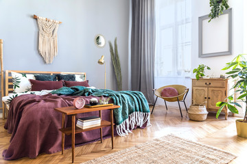 Stylish and luxury interior of bedroom with design furnitures, honey yellow armchair, beige macrame, gold lamp and elegant accessories. Beautiful bed sheets, blankets and pillows. Warm home decor.