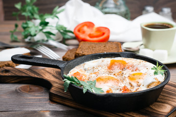 Ready-to-eat breakfast: shakshuka from fried eggs with tomatoes and parsley in a pan, bread with butter and coffeee on a wooden table