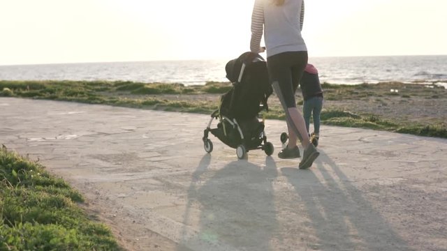 An athletic woman runs with a pram. Next to her rides her little daughter on a scooter. They run along the promenade by the sea