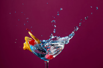 Colorful cocktail in glass with splashes garnished with lemon and cherries.