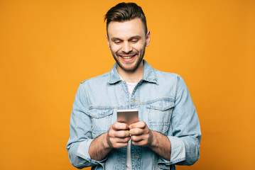 Young modern stylish man in denim is using smartphone in his hands while standing over yellow...