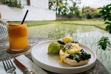 Close up Healthy Breakfast with Bread Toast and Poached Egg with spinach, avocado on wooden table with view on rice field. Orange smoothie on the background.