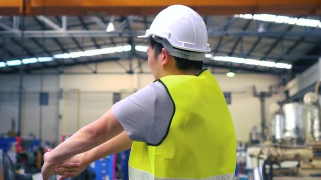 Back view of Asian industrial engineer in helmet and waistcoat doing stretching exercises while relaxing during break on factory