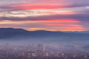 Unreal, purple sunset sky above night cityscape with city lights covered by thick mist