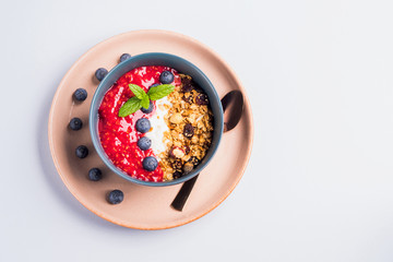 Yogurt smoothie bowl with blended raspberries, blueberries and oat matcha granola on pastel background. Delicious healthy breakfast concept