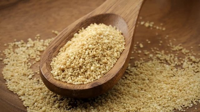 Heap of raw, uncooked couscous in wooden scoop on wood plate rotating