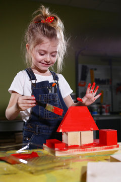 The little girl in the carpentry workshop paints with red paint wooden rectangles, triangle panes, cubes.Head down, not looking at the camera. . The concept of children's learning.