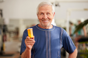 Smiling senior man holding bottle of pills. Portrait of older man looking at camera on blurred background. Sport supplement concept.