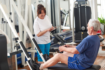 Cheerful senior couple at gym. Mature woman with digital tablet and man exercising on machine at fitness club. People, sport, training and modern technology concept.