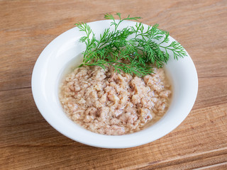White porcelain bowl with shredded tuna in oil for salad and green dill on a wooden background. Seafood, healthy eating and cook at home.