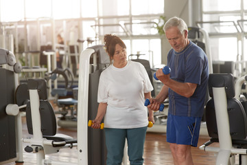 Elderly couple working out at gym. Senior man lifting dumbbells at fitness center. Sport activity for seniors.