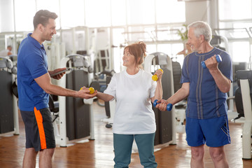 Personal trainer helping lift dumbbells to senior woman. Happy elderly couple lifting dumbbells while male instructor guiding them. Easy strength training workouts.