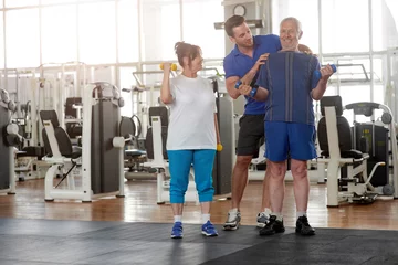  Personal trainer helping lift dumbbells to seniors. Elderly man lifting weights at fitness center with personal trainer. People, retirement, sport, active lifestyle. © DenisProduction.com