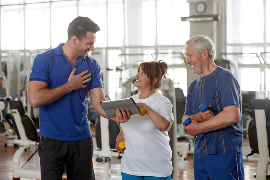 Personal male trainer showing something on digital tablet. Happy senior couple at gym. Sport, fitness, technology, lifestyle and people concept. - Powered by Adobe