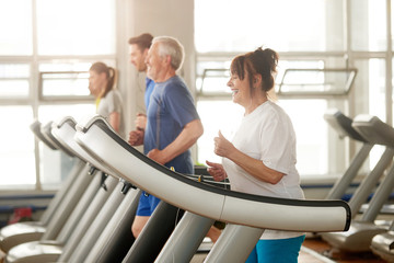 Mature caucasian woman running on treadmill. Happy retired woman listening to music during training in fitness club. Sport gives energy.