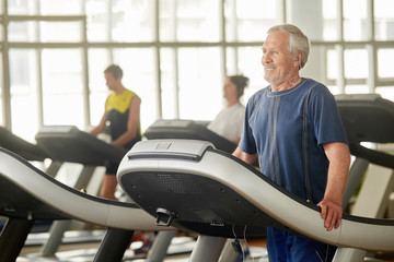 An elderly man is engaged in fitness. Happy senior man in eraphones listening to music while exercising on treadmill in gym.