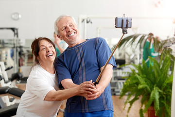 Fototapeta premium Smiling elderly couple taking selfie at gym. Beautiful senior people resting in gym taking selfie with monopod. People, sport, modern technology and leisure concept.