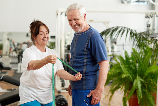 Beautiful elderly couple at sport club. Happy senior lady with measuring tape showing her fitness results at fitness club. People, sport, diet and weight loss concept.